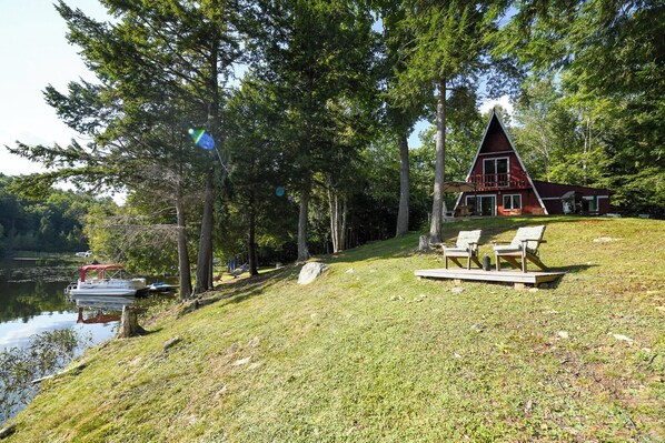 Una cabaña junto al lago con techo rojo, rodeada de árboles, con mobiliario exterior y un muelle.