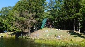 A cabin with a green roof, surrounded by dense forest, near a calm lake with a grassy area and a few chairs.
