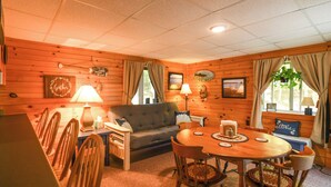 A cozy cabin-style room with a wooden interior, a sofa, a dining table with chairs, and a small kitchen area.
