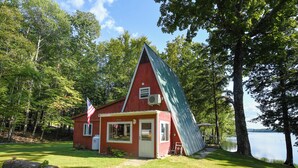 A red cabin with a slanted roof, surrounded by trees and a lake.