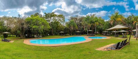 A swimming pool with two separate sections, surrounded by grass and trees. There are lounge chairs and umbrellas around the pool area.