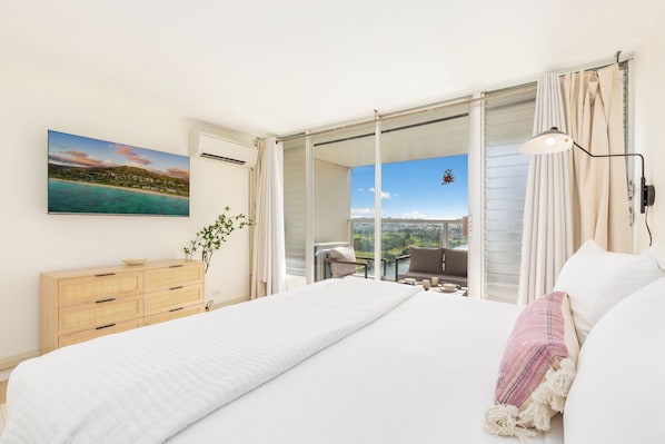 A bedroom with a large bed, a wooden dresser, a plant, and a balcony with a view of the sea.
