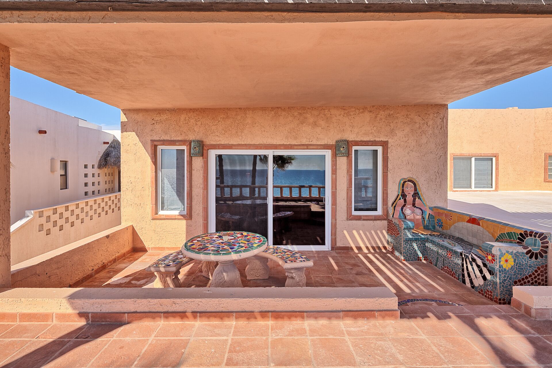 A covered outdoor area with a patio table and chairs, a mosaic bench, and a view of the sea through a glass door.