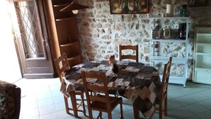 Un comedor con una mesa, sillas, una pared de piedra, un cuadro y una estantería con utensilios de cocina.