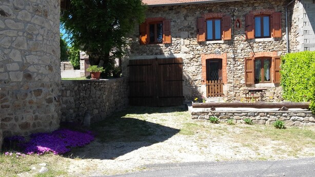 Un edificio de piedra con contraventanas de madera, una entrada protegida por una reja y un pequeño espacio exterior con mesas y sillas.