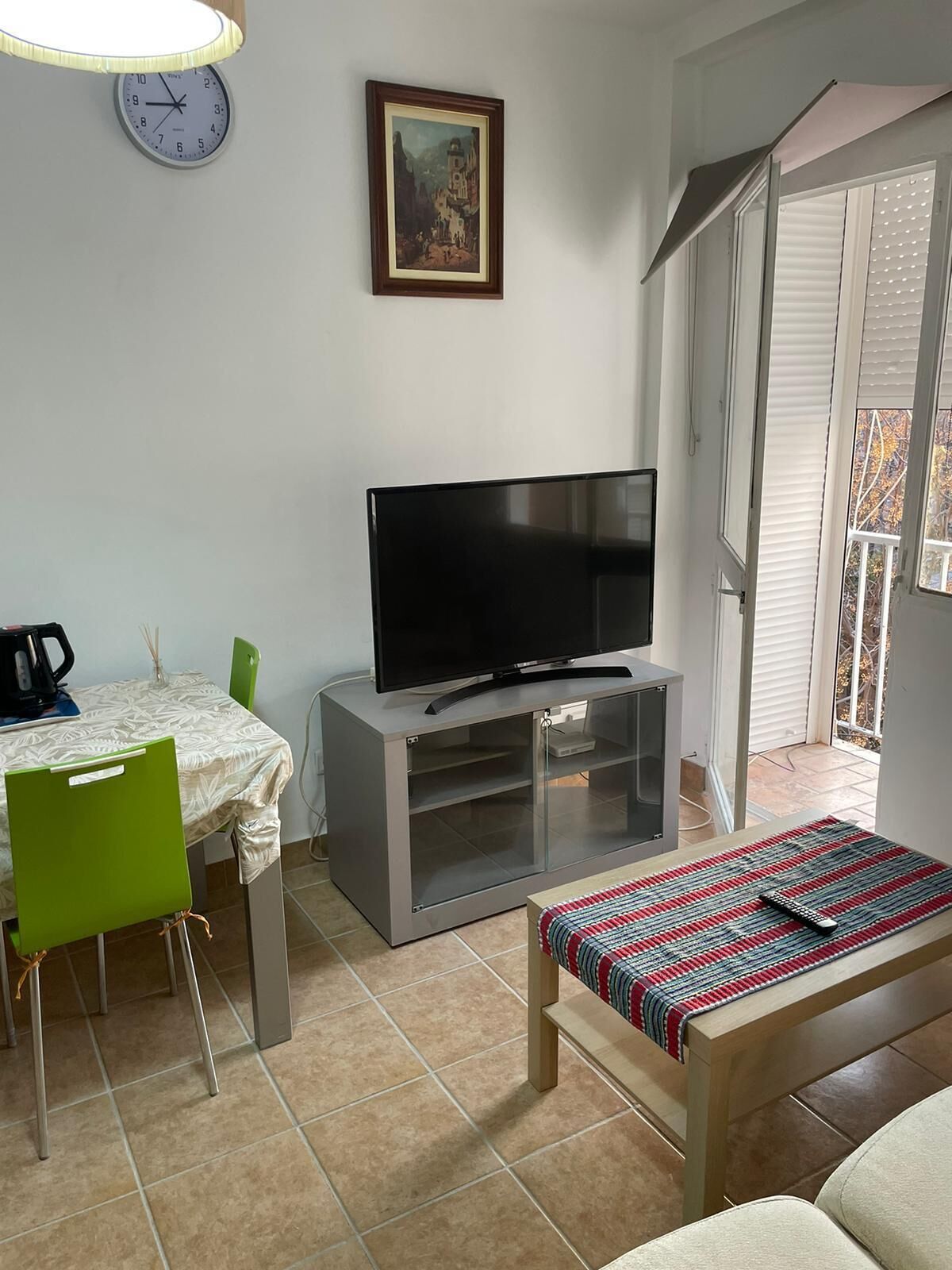 Un salon avec un téléviseur à écran plat posé sur un meuble, une table à manger avec des chaises, une horloge au mur et une fenêtre avec des stores.