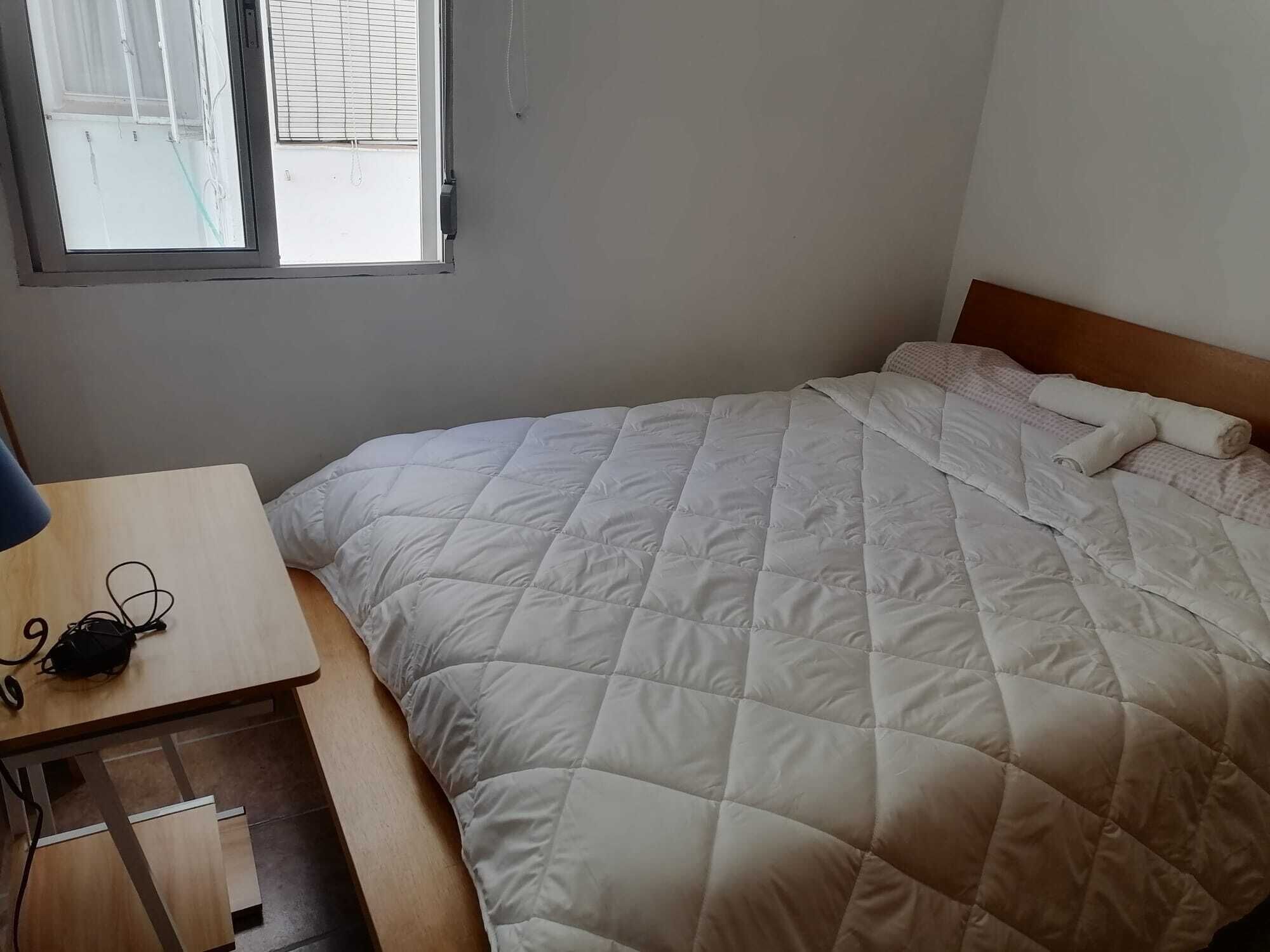 Een net opgemaakt bed met een gewatteerd dekbed, een houten nachtkastje met een lamp en een raam met jaloezieën.