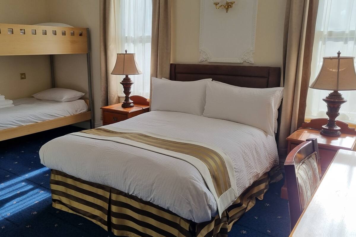 Ein Hotelzimmer mit Etagenbett, einem Einzelbett, zwei Nachttischen mit Lampen und einem großen Fenster mit Vorhängen.