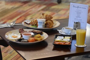 Ein Frühstückstisch mit Tellern mit Eiern, Speck, Toast und Gebäck, einer Speisekarte, einer Kaffeetasse und einem Glas Orangensaft.