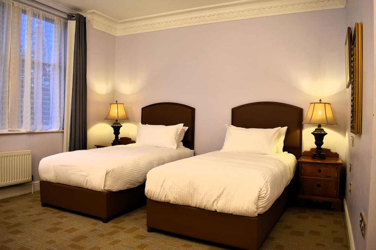 Ein Hotelzimmer mit zwei Betten, einem Fenster mit Vorhängen, einem Heizkörper und zwei Nachttischlampen.