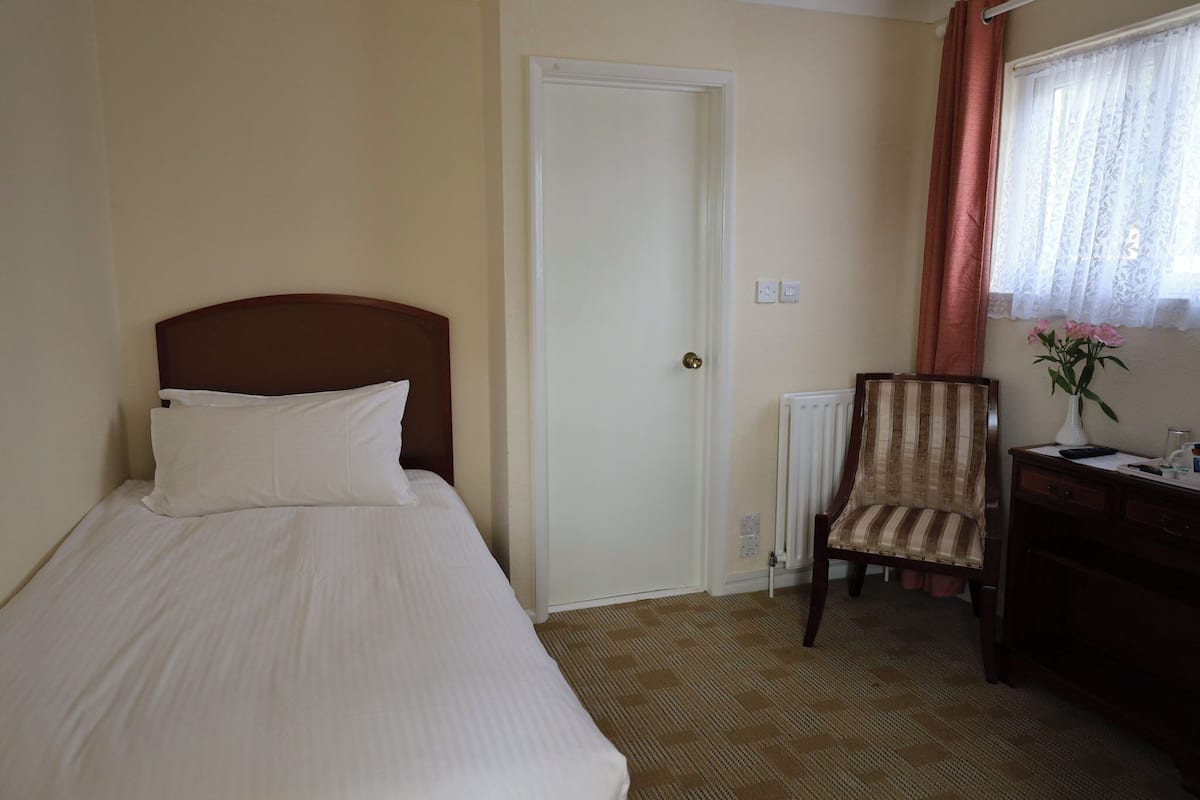 Ein Hotelzimmer mit Bett, Sessel, Schreibtisch, Heizkörper und einem Fenster mit Vorhängen.