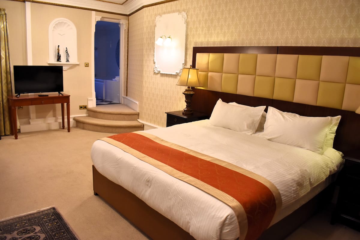 Ein Hotelzimmer mit einem großen Bett, einem Fernseher auf einem Holztisch, einem Nachttisch mit Lampe und einer kleinen Treppe zu einem Badezimmer.