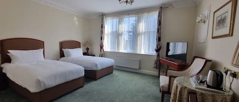 Habitación de hotel con dos camas individuales, un escritorio con televisor, un radiador y una ventana con cortinas.