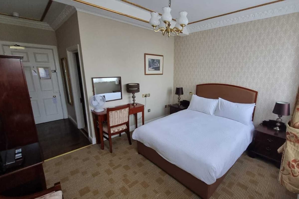 Ein Hotelzimmer mit einem großen Bett, einem Schreibtisch mit Stuhl, einem Nachttisch mit Lampe, einem Spiegel und einer Chandelier.