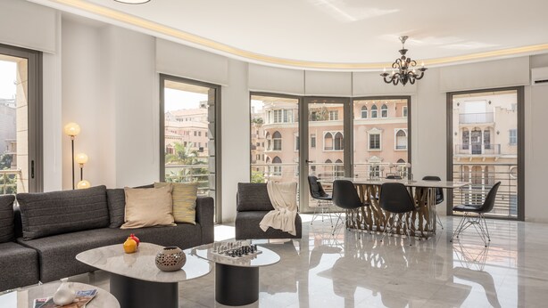 Uma sala moderna com sofá, mesa de centro e área de jantar. Janelas amplas oferecem vista para edifícios e uma varanda.