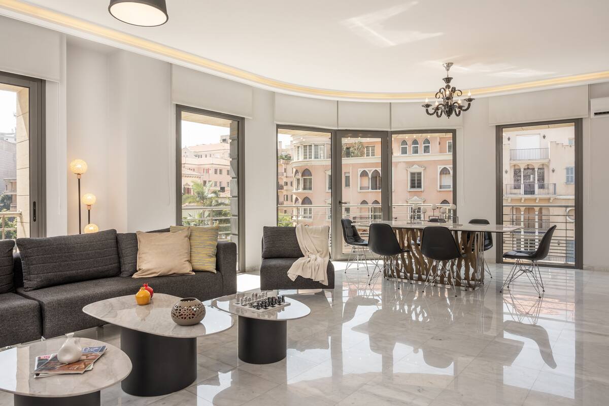 A modern living room with a sofa, coffee table, and dining area. Large windows offer views of buildings and a balcony.