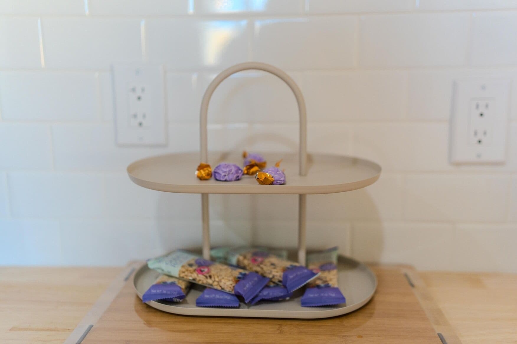 A two-tiered metal stand with a handle, holding purple and gold wrapped chocolates on a kitchen counter.