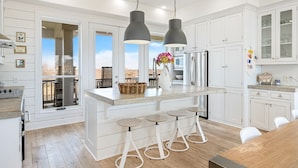 A modern kitchen with white cabinetry, a central island with stools, pendant lighting, and a view through large windows. - #Beach Please - Amazing Views From All Directions - Close to the beach (Crystal Beach)