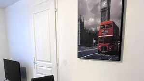 Une pièce moderne avec une table blanche et des chaises noires, une porte blanche et un tableau représentant un autobus à deux étages rouge avec un beffroi.
