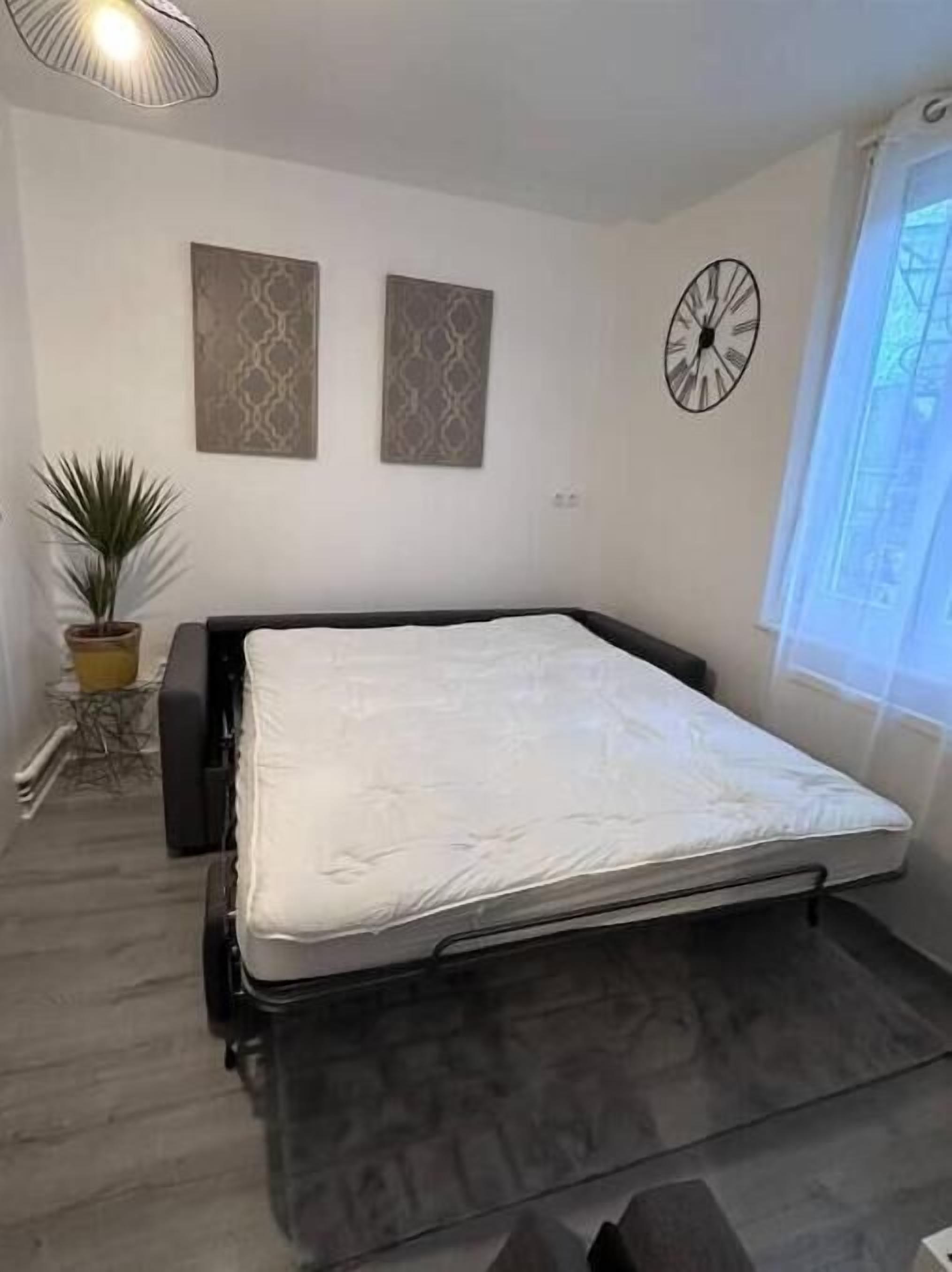 A bedroom with a bed, a potted plant, two wall art pieces, a clock, and a window.