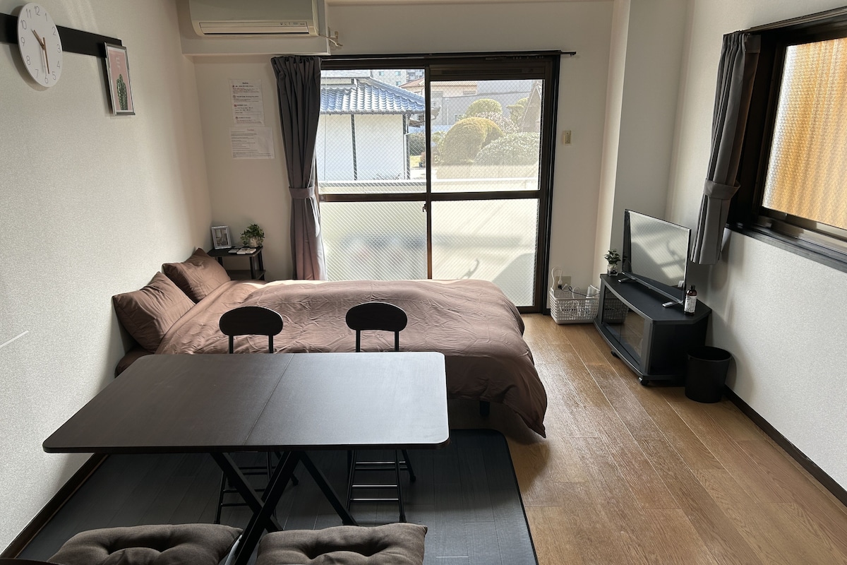 A compact room with a bed, a small table, two chairs, a TV, and a window with a view of buildings.