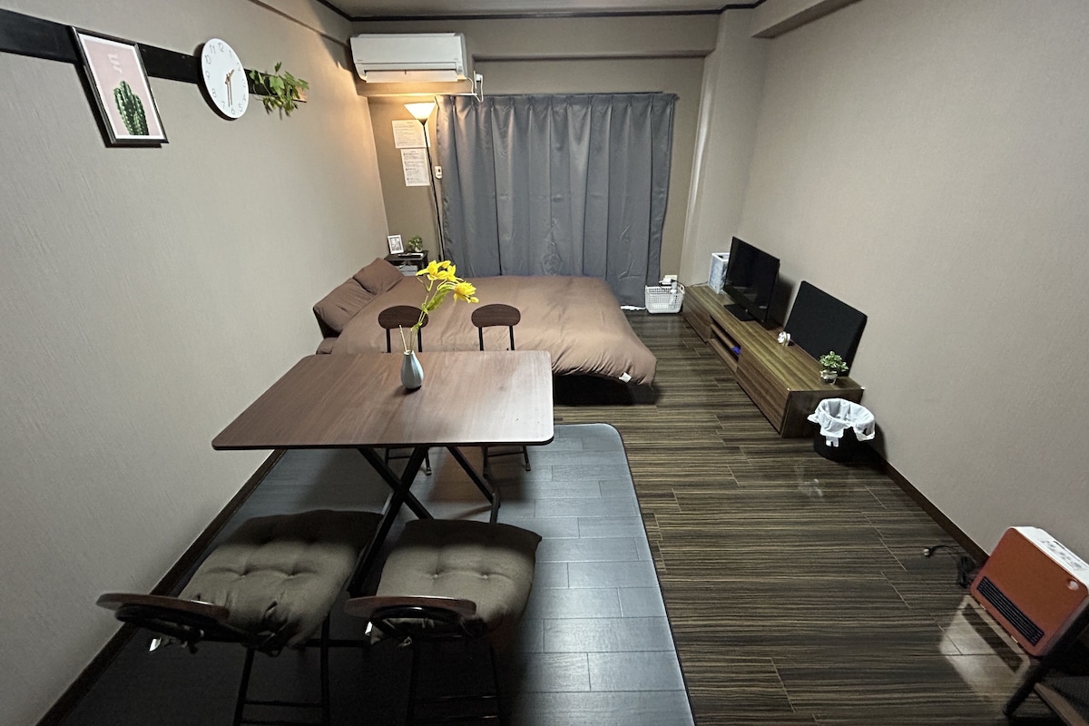 A compact hotel room with a bed, a small dining table with chairs, a TV, and a bedside table with a lamp.