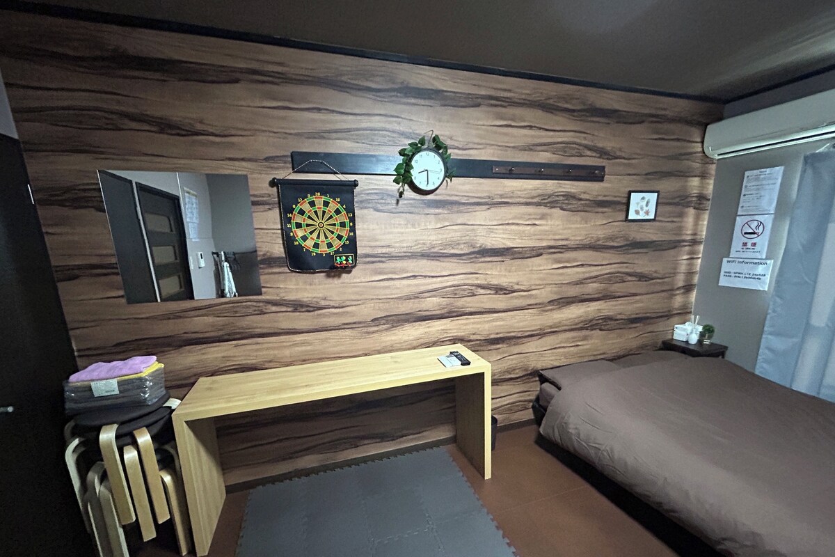 A room with a wooden wall, a dartboard, a bed, and a desk with a chair.
