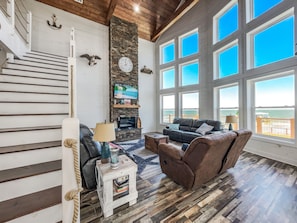 A spacious living room with a fireplace, a large TV, and a view of the ocean through large windows. - 2 Lodge Combo - Sleeps 30 w/Pool + Private Pier (San Leon)