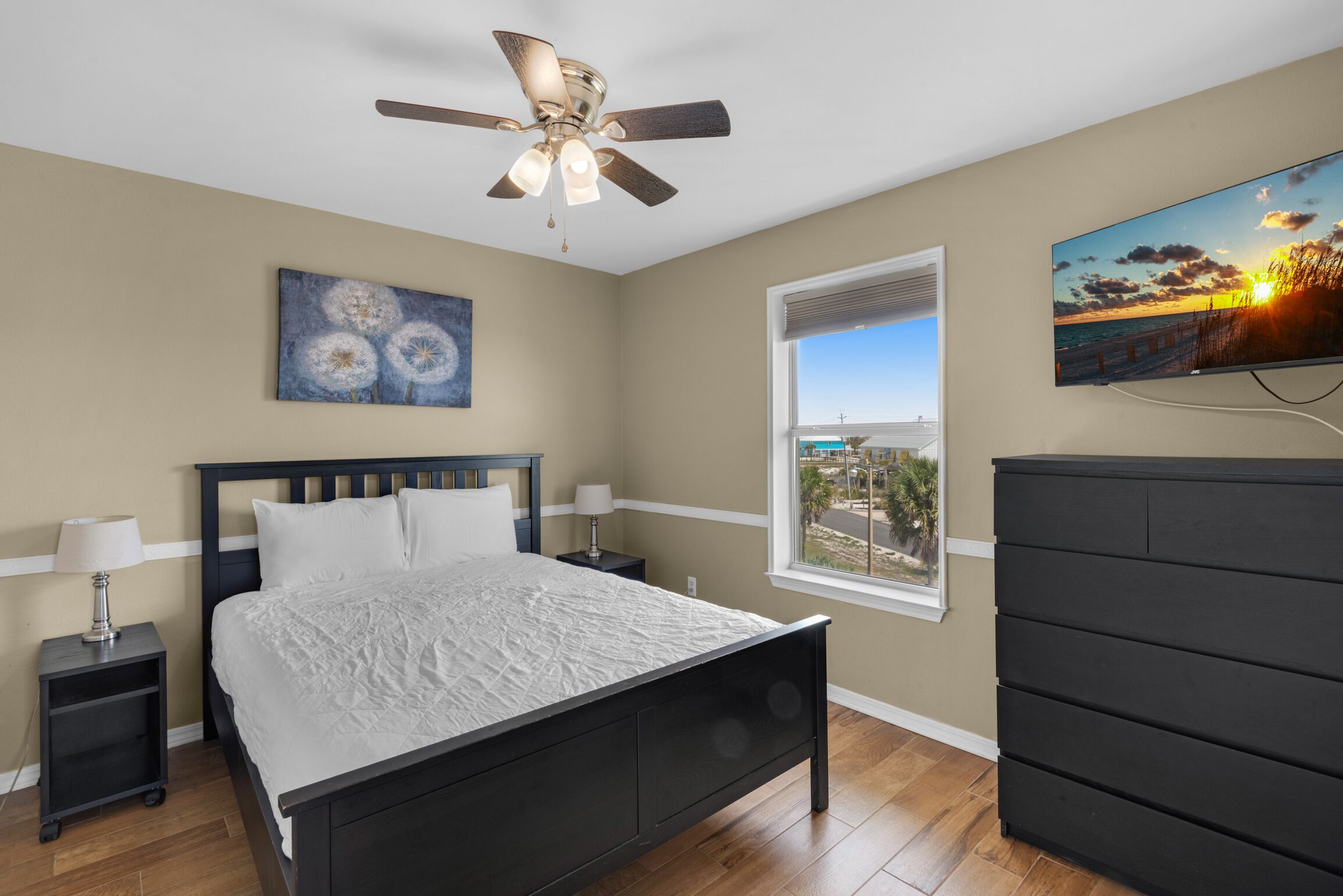 A bedroom with a bed, a ceiling fan, a window with a view, a painting, and a TV.