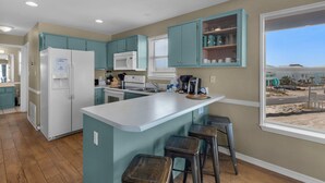 Une cuisine avec des armoires de couleur turquoise, un réfrigérateur blanc, un four à micro-ondes et une grande fenêtre donnant sur une plage et des immeubles.