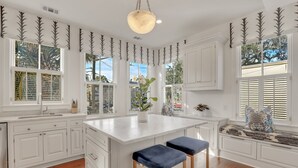 Une cuisine avec des meubles blancs, un îlot central doté de tabourets et de grandes fenêtres équipées de volets.