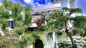 A white building with a balcony, an arched entrance, and a small garden area with various plants.