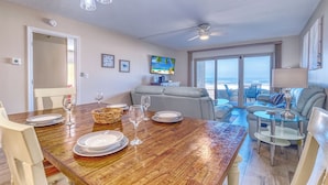 A dining area with a wooden table set for four, a grey sofa, and a view of the ocean through large windows. - Elevated Ground Level Direct Gulf Front 2 Bedroom/2 Bath Condo (Redington Shores)