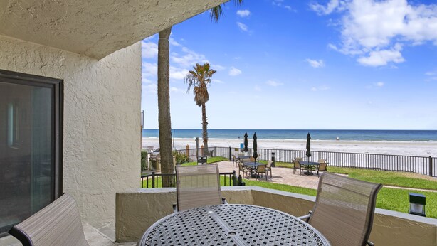 A balcony with a table and chairs, overlooking a beach with palm trees and a fence. - Elevated Ground Level Direct Gulf Front 2 Bedroom/2 Bath Condo (Redington Shores)