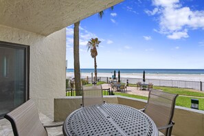 A balcony with a table and chairs, overlooking a beach with palm trees and a fence. - Elevated Ground Level Direct Gulf Front 2 Bedroom/2 Bath Condo (Redington Shores)