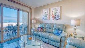 A living room with a glass coffee table, a leather sofa, and a view of the beach through large sliding glass doors. - Elevated Ground Level Direct Gulf Front 2 Bedroom/2 Bath Condo (Redington Shores)