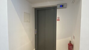 A narrow hallway with a wooden floor, a closed elevator door, a fire extinguisher mounted on the wall, and a digital display above the elevator. - Heart of Nottingham - turret studio (Nottingham)