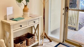 Una habitación con un escritorio blanco, dos cuadros enmarcados, un florero con una planta, un perchero y un ventanal corredero que da al exterior.