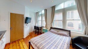A bedroom with a bed, a desk, a chair, a TV mounted on the wall, and a window with curtains. - Studio with Great City Views (Nottingham)
