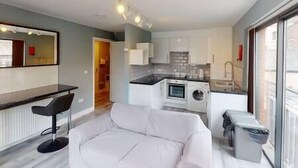 A modern kitchen with a black countertop, a built-in oven, and a washing machine. There is a white sofa in the living area and a bar counter with two stools. - Lovely 2 bedroom flat with free parking (Nottingham)