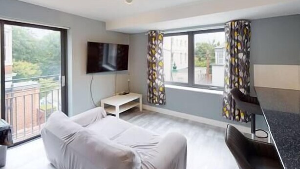 A modern living room with a sofa, a flat-screen TV mounted on the wall, and a balcony with patterned curtains. - Lovely 2 bedroom flat with free parking (Nottingham)