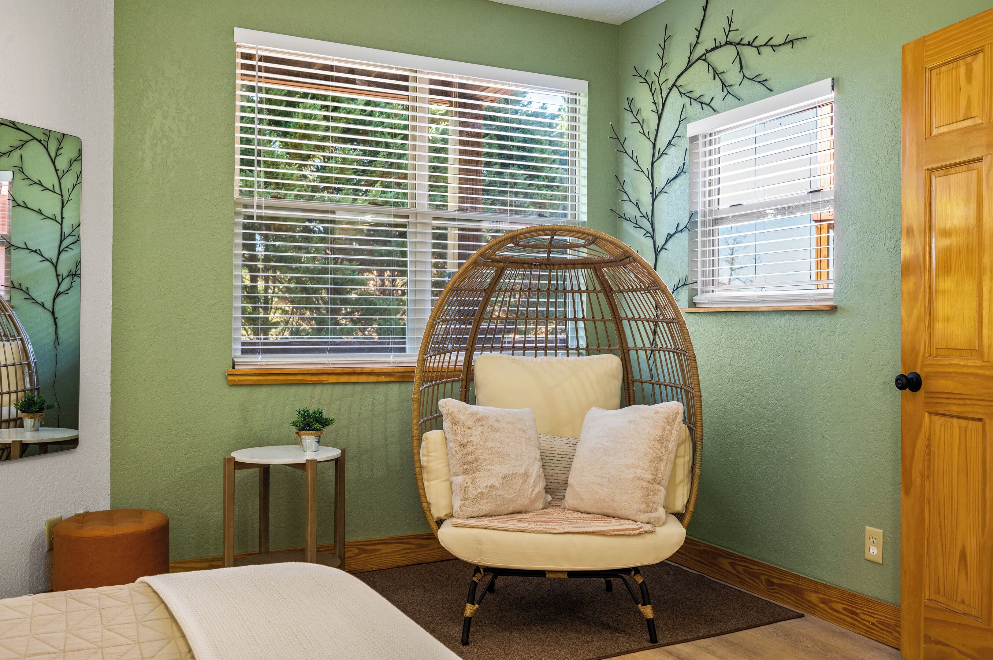 A room with a wicker chair, a small table with a plant, and two windows with blinds.