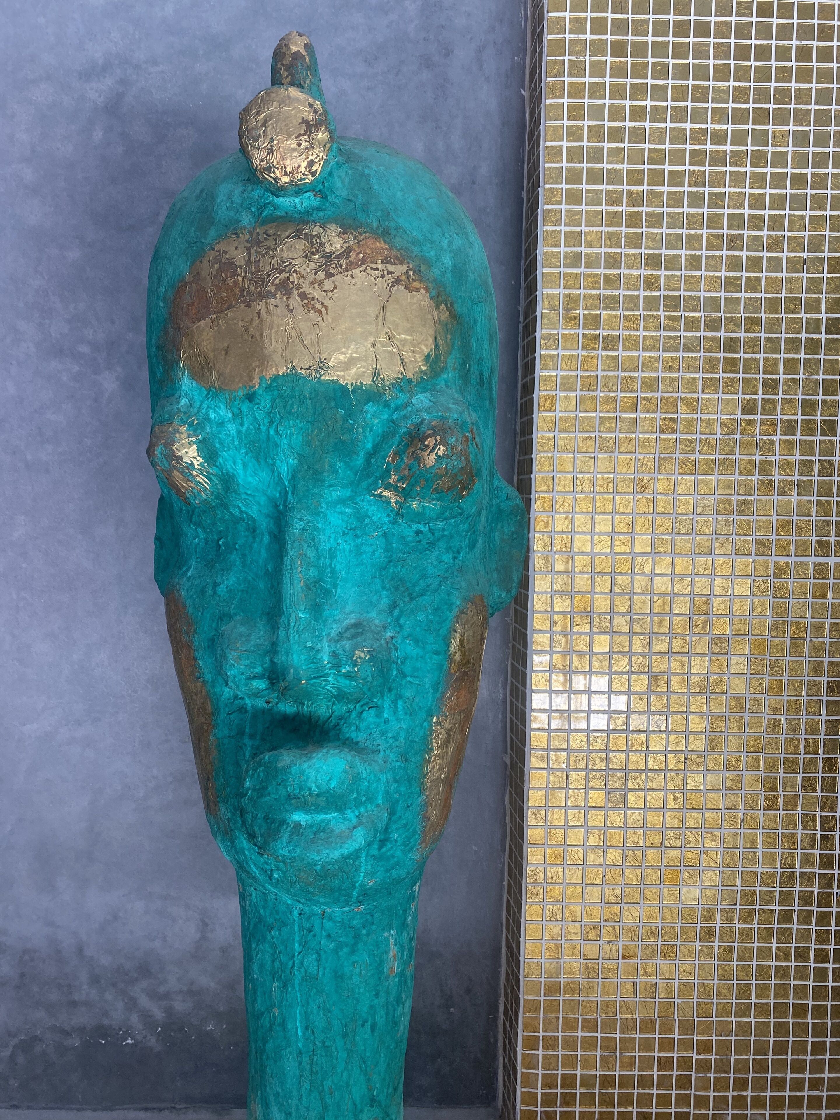 A sculpture with a teal and gold color scheme, placed against a grey wall with a gold-tiled section to its right.