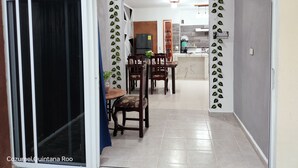 A dining area with a table and chairs, a kitchen in the background, and a decorative leaf pattern on the walls.
