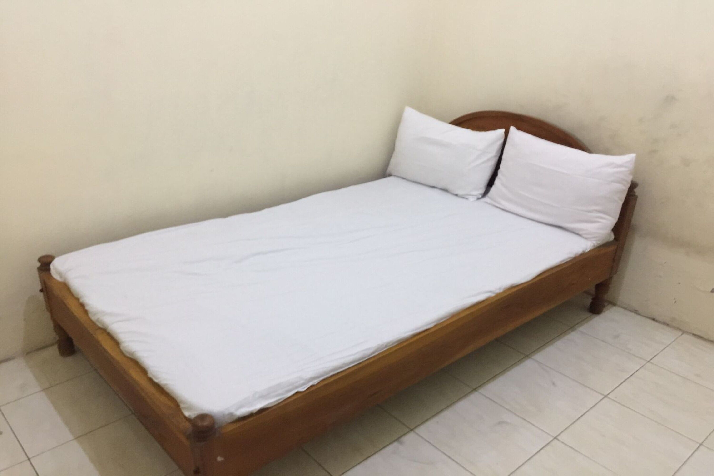 A single bed with a wooden frame and white bedding in a room with plain walls and tiled flooring.