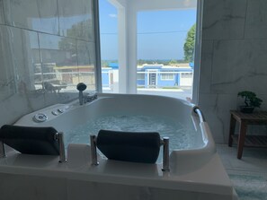 A modern bathroom with a large bathtub, a view of the outside, and a small wooden table with a potted plant.