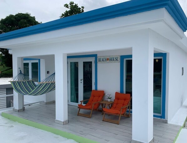 A beach house with a sign reading "BEACH HOUSE," two orange lounge chairs, and a hanging chair on a covered patio.