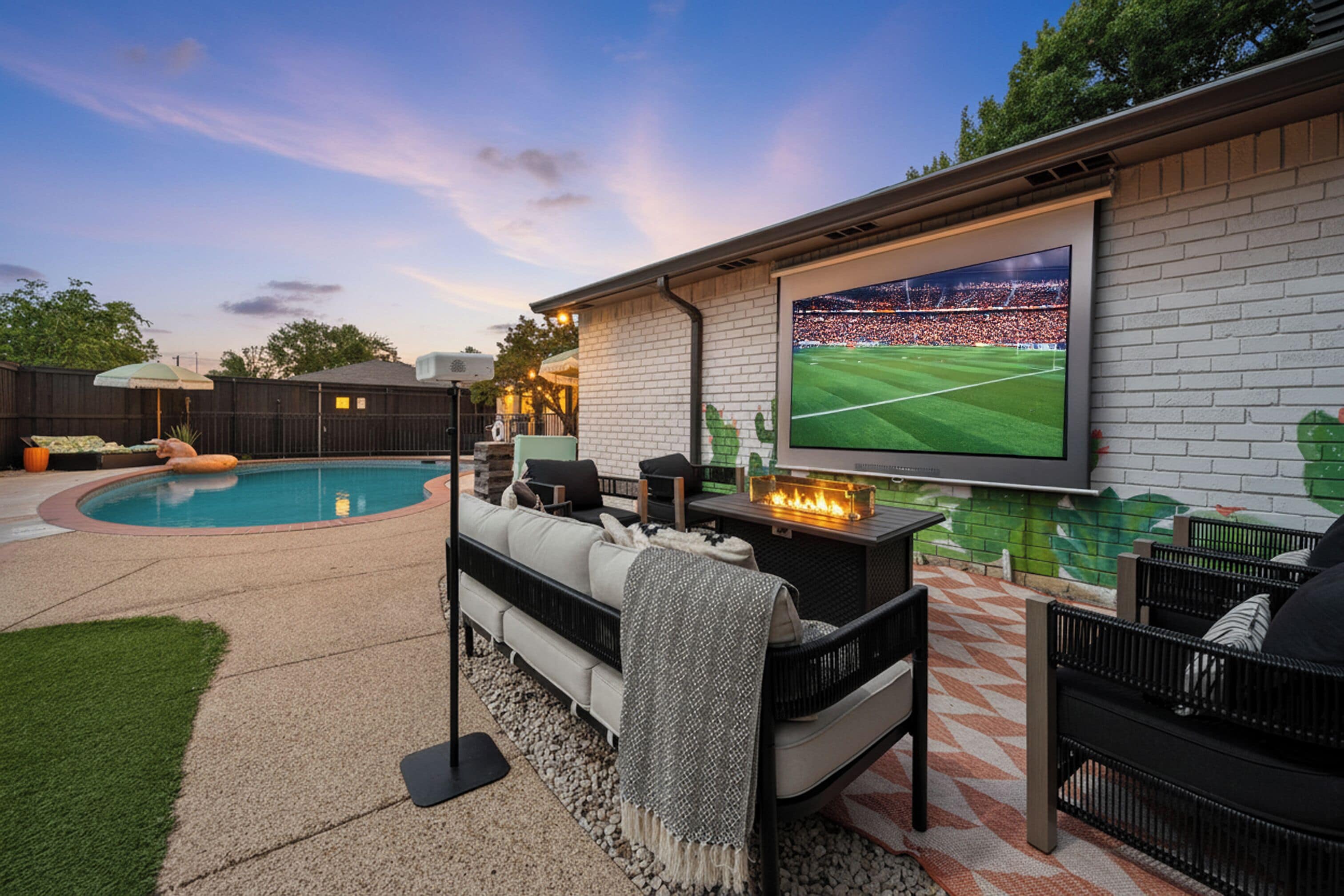 A backyard with a pool, outdoor seating, a fire pit, and a large TV screen displaying a sports event.