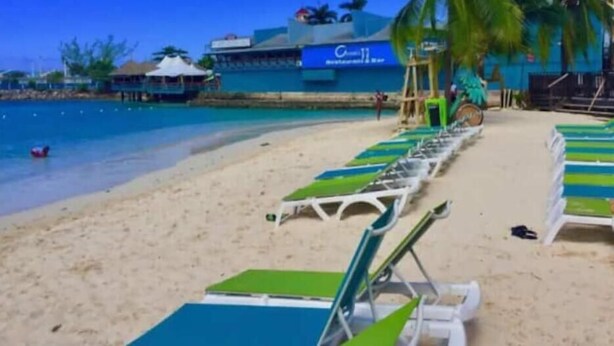Una spiaggia con file di lettini verdi e bianchi, un cielo azzurro limpido e un edificio con un cartello che recita 'Coco II'.
