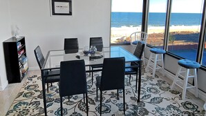 Un coin repas avec une table et des chaises en verre, un ventilateur de plafond, une bibliothèque et une grande fenêtre donnant sur l’océan.